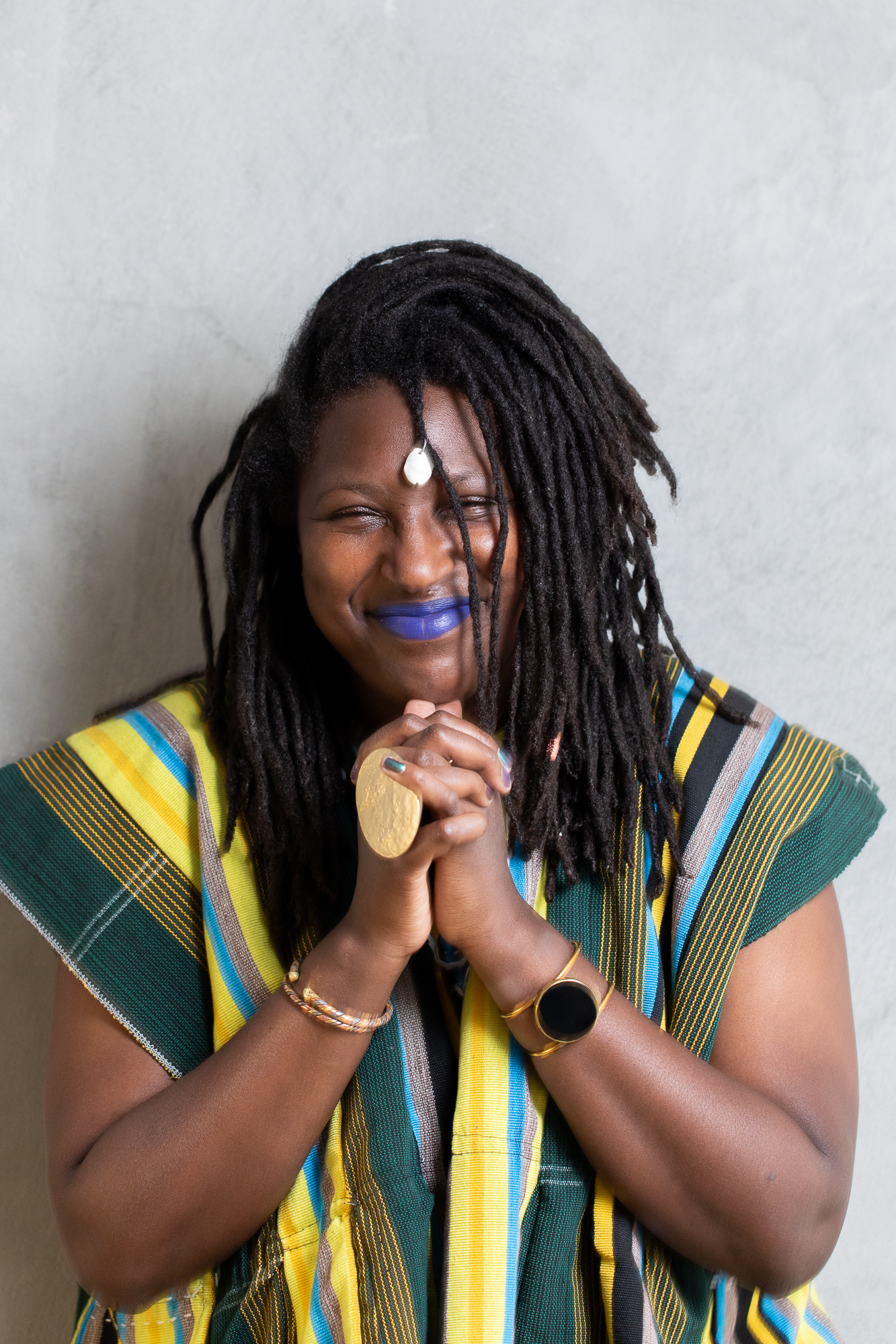 PHOTO DE MOI GRANDE FEMME NOIRE SOURIANT DEVANT UN MUR GRIS J'AI UNE VESTE A RAYURES JAUNES BLEUES ET VERTES ET UNE GROSSE BAGUE EN FORME DE LUNE, DES LOCKS MI-LONGUES AVEC UNE RAIE SUR LE COTÉ.Elle porte un rouge à lèvres bleu.